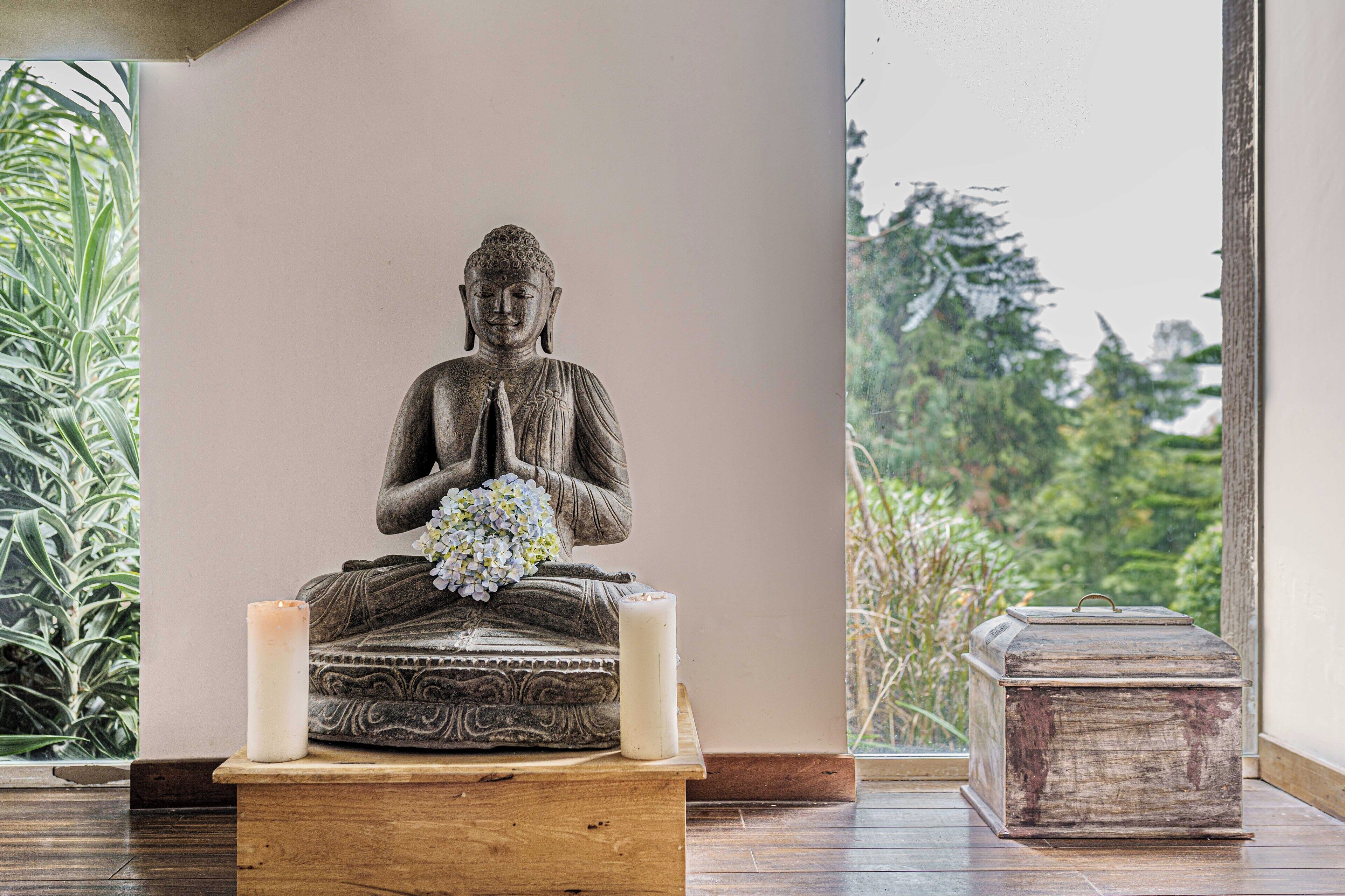 Una estatua de Buda sobre un pedestal, con una vela y flores, ubicada en una habitación que ofrece vistas a la vegetación a través de una ventana.