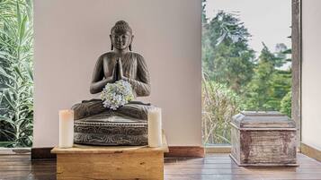 Una estatua de Buda sobre un pedestal, con una vela y flores, ubicada en una habitación que ofrece vistas a la vegetación a través de una ventana.