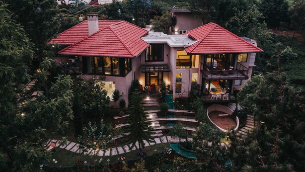 Una casa grande y moderna con dos secciones de techo rojo, una entrada central y un jardín ajardinado con un sendero sinuoso.