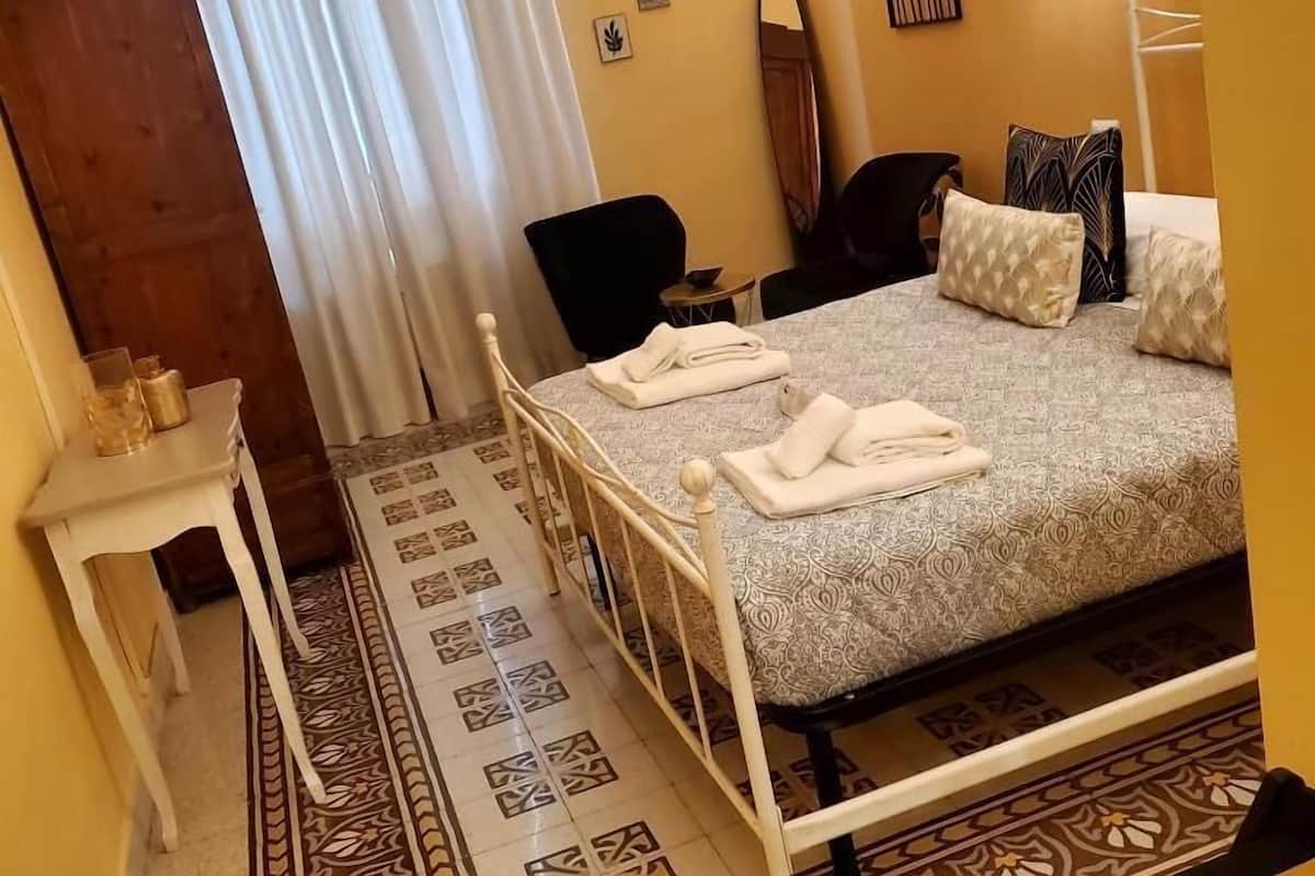 A bedroom with a bed, a chair, a small table with a glass and a bottle, and a door.