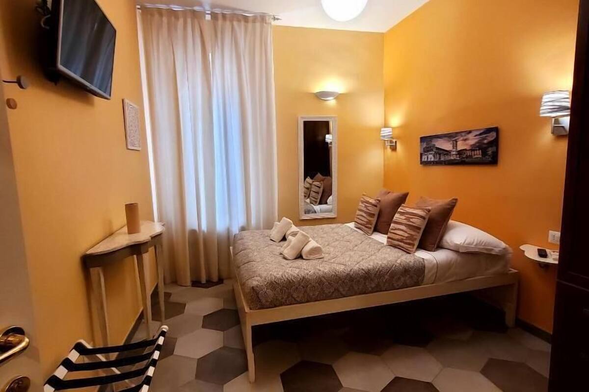 A compact hotel room with a bed, a television mounted on the wall, a small table, and a mirror.