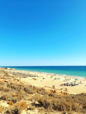 Ein Strand mit Menschen und Sonnenschirmen, sandigem Untergrund und blauem Himmel.