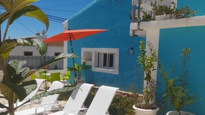 Een blauw gebouw met witte muren, een klein raam en een rode parasol. Er staan witte lounge stoelen en potplanten.
