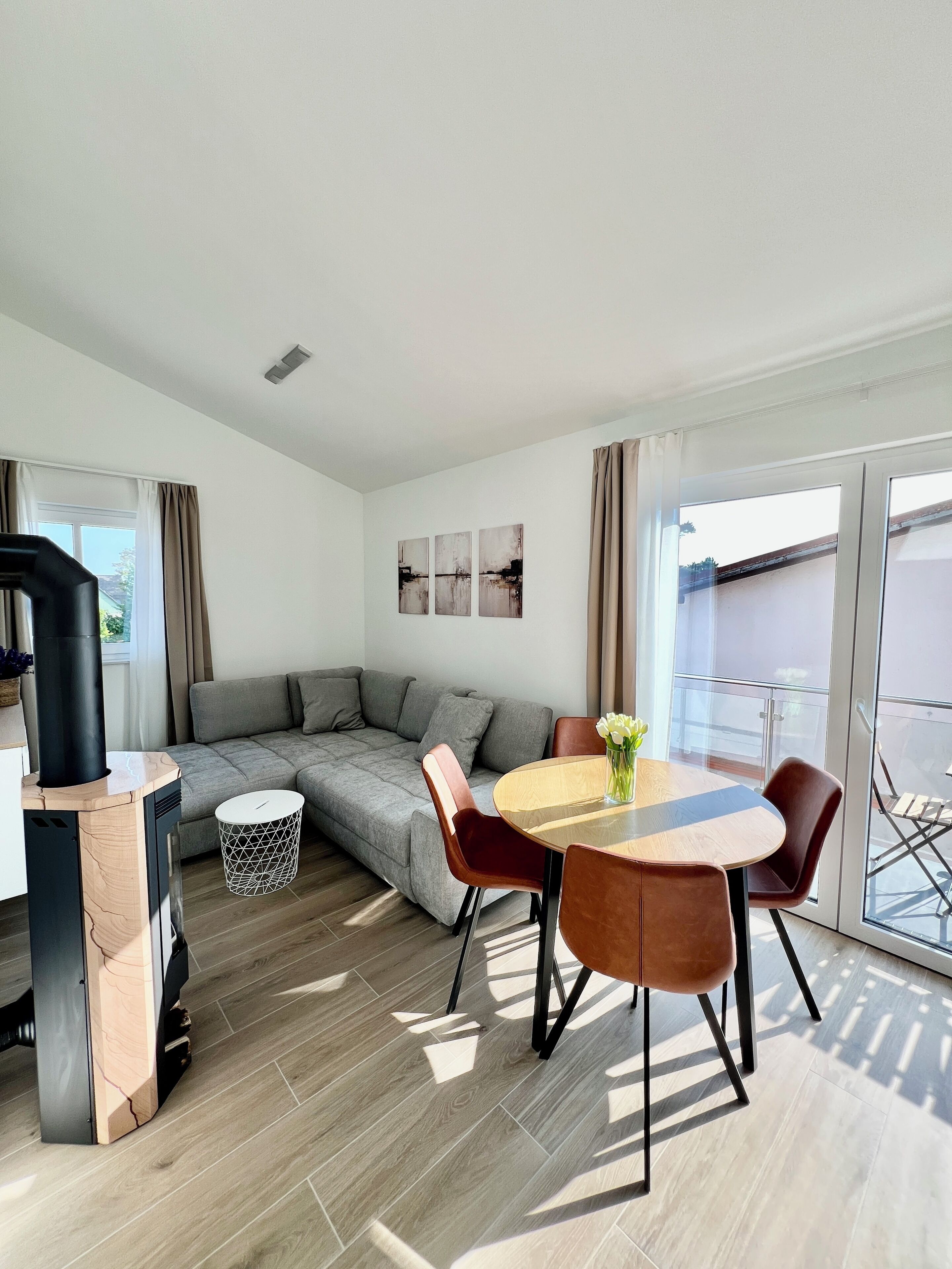 A modern living room with a grey sofa, a round wooden table with chairs, a black stove, and a glass door leading to a balcony.