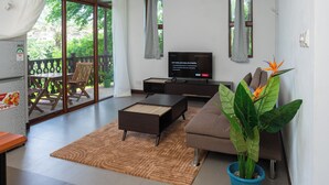 A modern living room with a sofa, a coffee table, and a TV. There is a balcony with a wooden table and chairs.