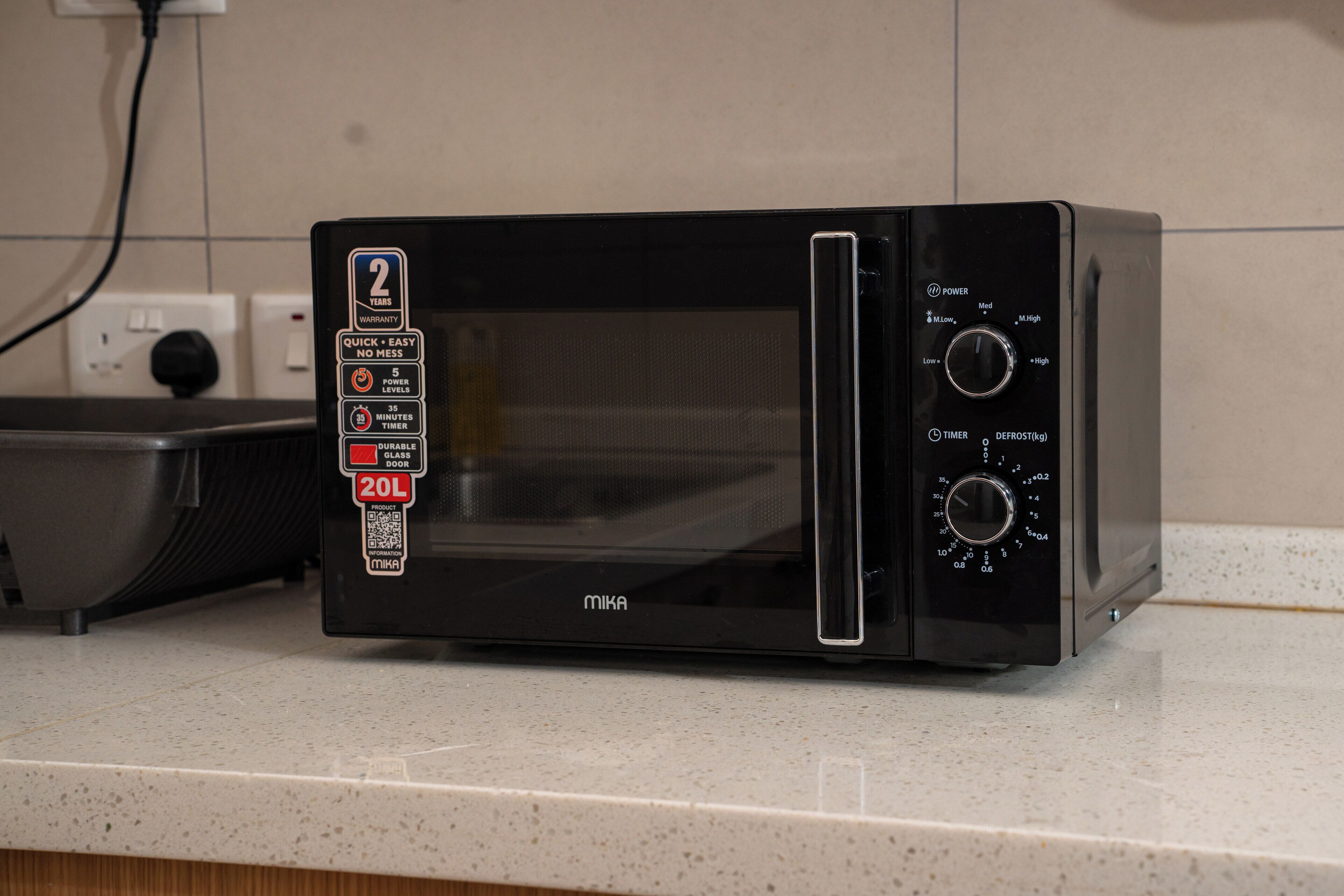 A black microwave oven with a glass door, a power dial, a timer dial, and a defrost setting. It is placed on a kitchen countertop next to a sink and a power outlet.