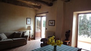 A cozy living room with a sofa, a table with a checkered tablecloth, and a vase of flowers. There is a lamp, a chair, and a view of the outside through a door.
