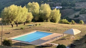 A rectangular swimming pool with a tiled deck, surrounded by grass and trees, with a hill and a house in the background.