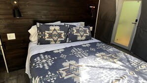 A bedroom with a bed, patterned bedding, a wall-mounted air conditioner, a bedside lamp, and a door to a balcony. -  Red Owl Retreat: The Peaks Geodesic Dome  (Bigfork)