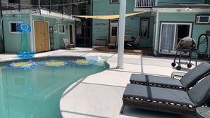 Zona de piscina con sillas de descanso, un porche cubierto y una casa al fondo.