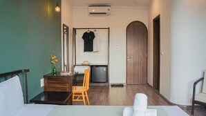 A hotel room with a bed, a small table with a chair, a desk with a chair, a wardrobe, and a door.