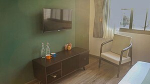 A room with a flat-screen TV mounted on the wall, a wooden cabinet with bottles and a cup, a chair, and a window with curtains.