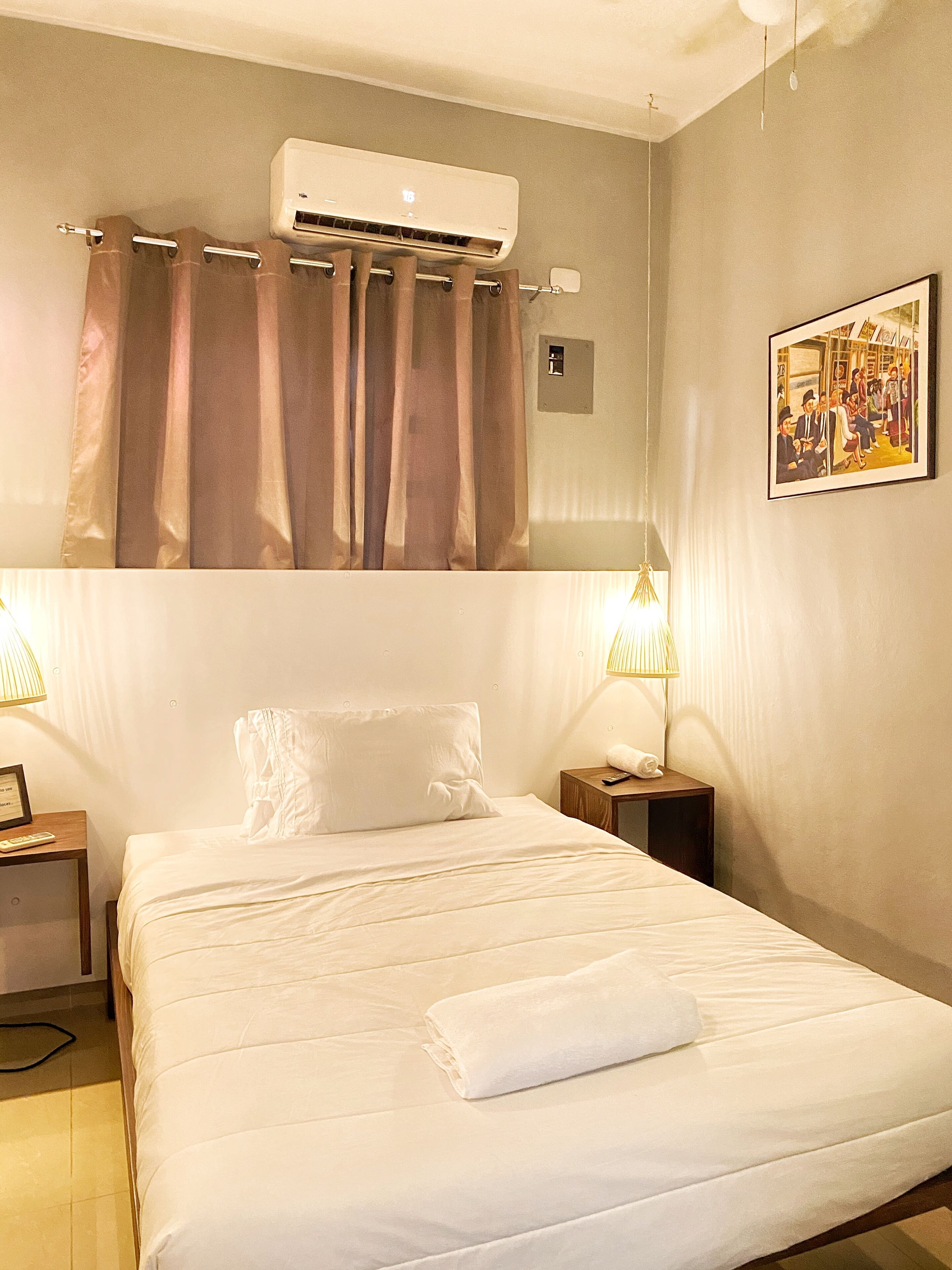 Quarto de hotel com cama, duas mesas de cabeceira, aparelho de ar condicionado, cortinas, um quadro na parede e um controle remoto sobre uma das mesas.