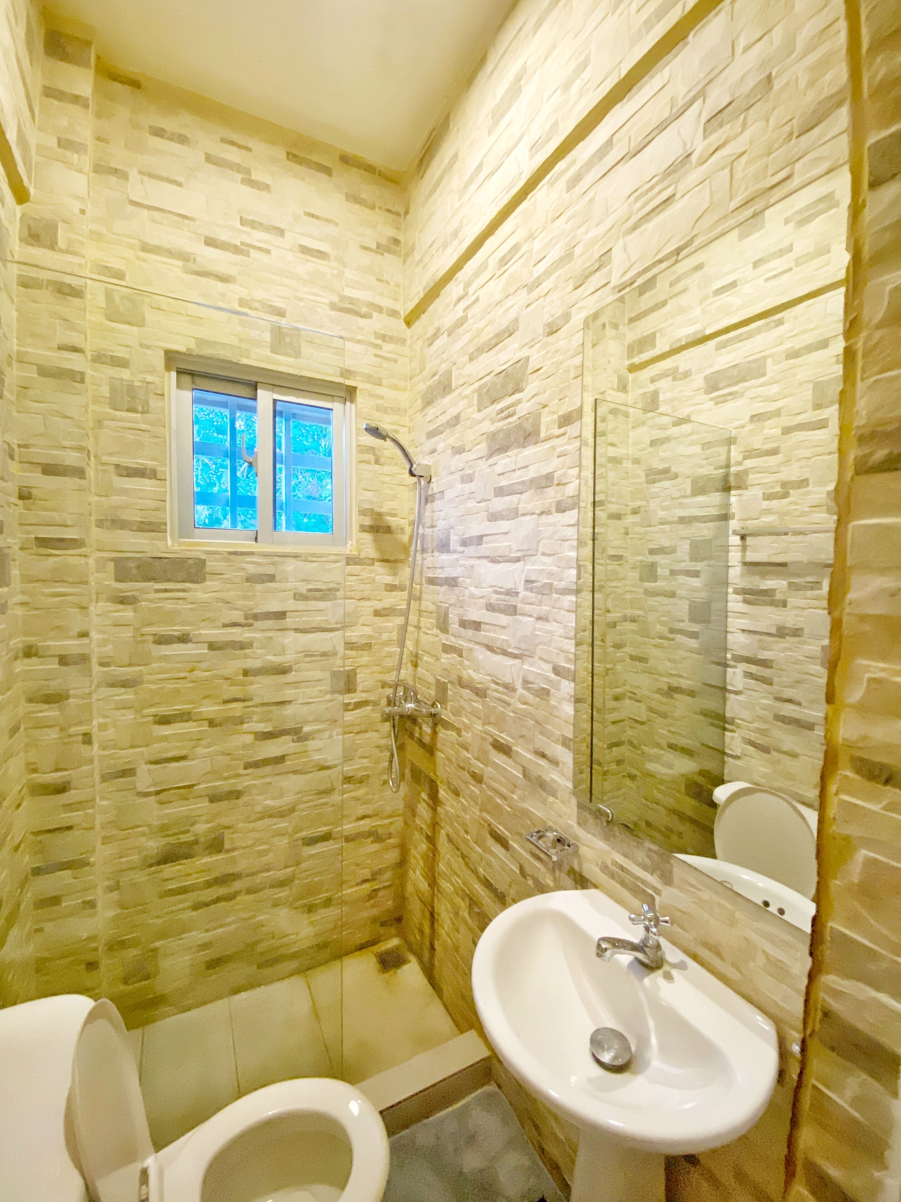 Une salle de bain avec un mur en pierre, une fenêtre, une douche, des toilettes et un lavabo.