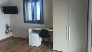 Una habitación con un armario blanco, un escritorio pequeño blanco, una silla negra, un televisor montado en la pared y una ventana con persianas.
