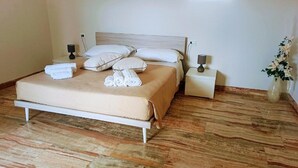 Una cama bien tendida con una colcha color beige y almohadas blancas, dos mesitas de noche con lámparas, y un jarrón con flores a la derecha.