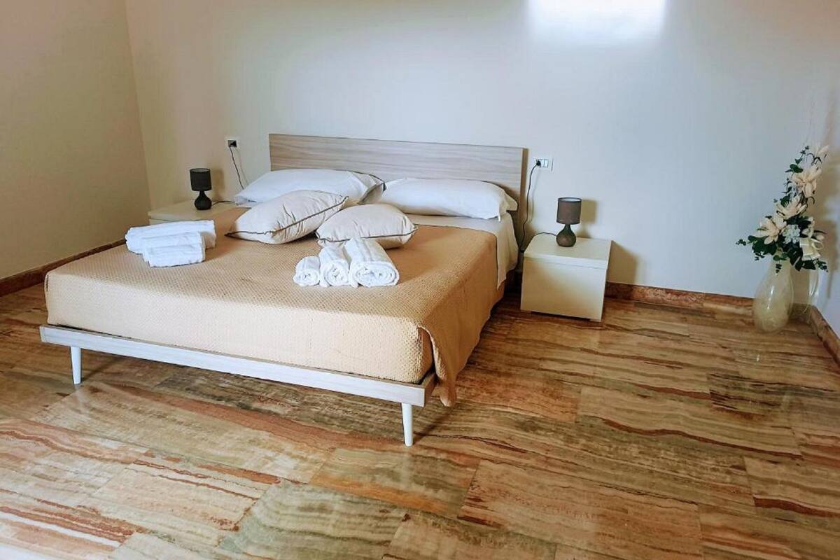A neatly made bed with a beige bedspread and white pillows, two bedside tables with lamps, and a vase with flowers on the right side.
