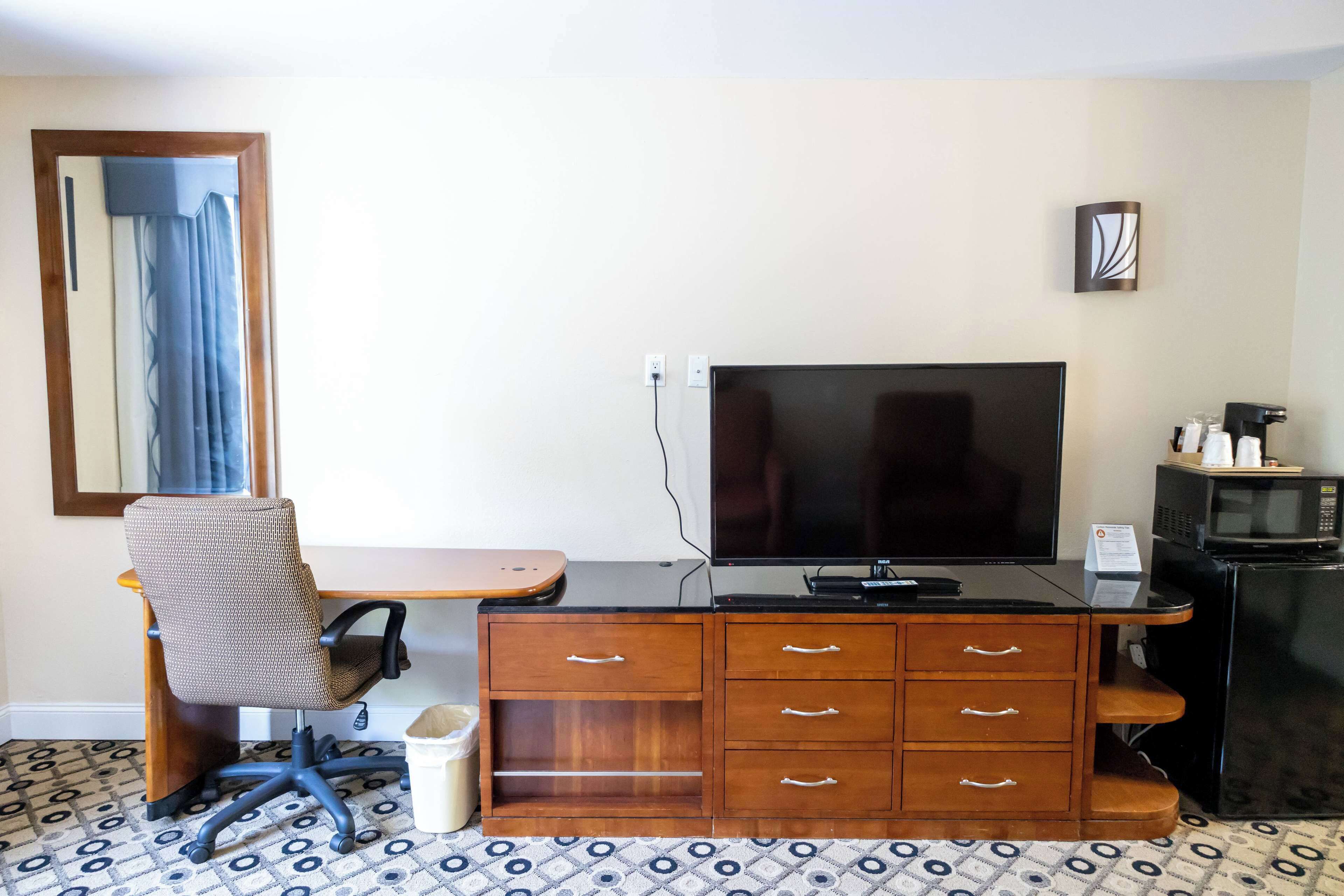 A hotel room with a wooden desk, a television, a microwave, and a trash bin.