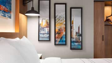 Ein Hotelzimmer mit einem Bett, weißen Kissen, einer schwarzen Lampe und drei gerahmten Stadtansichten an der Wand.