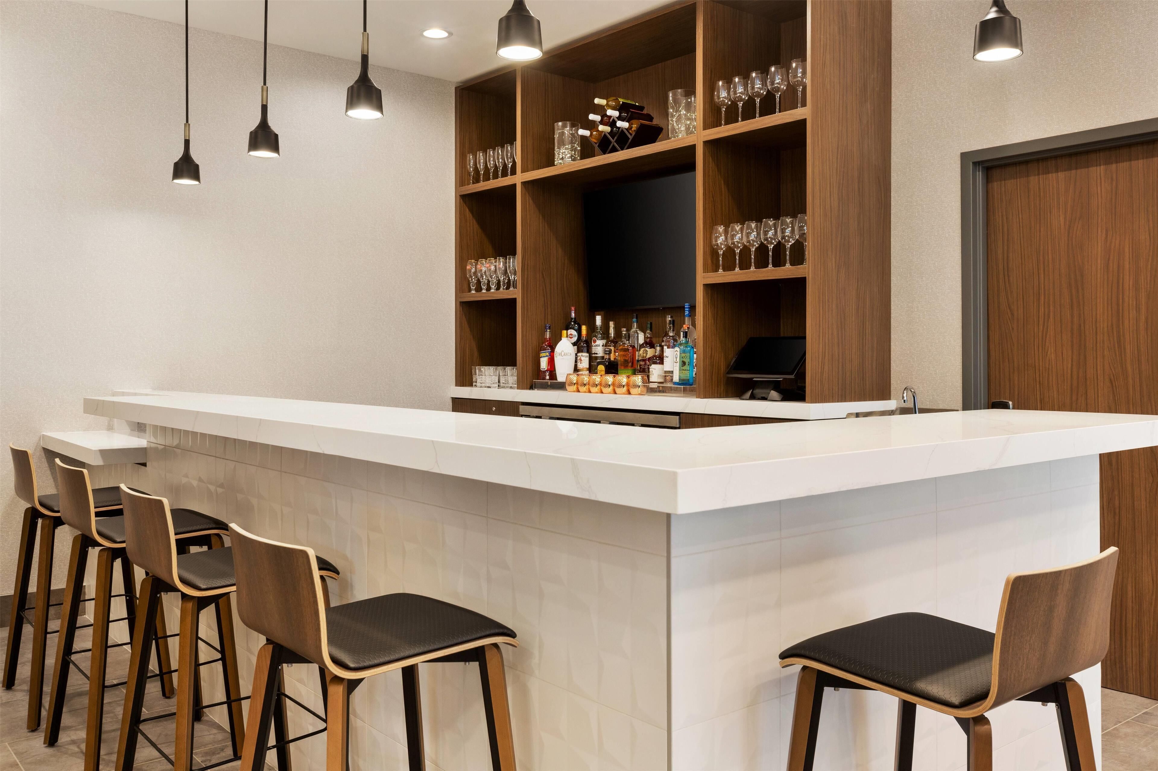 A bar area with a long counter, multiple stools, shelves with glasses and bottles, and hanging pendant lights.