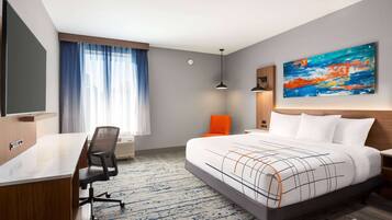 A modern hotel room with a large bed, a desk, and a chair. There is a painting on the wall and a window with curtains.