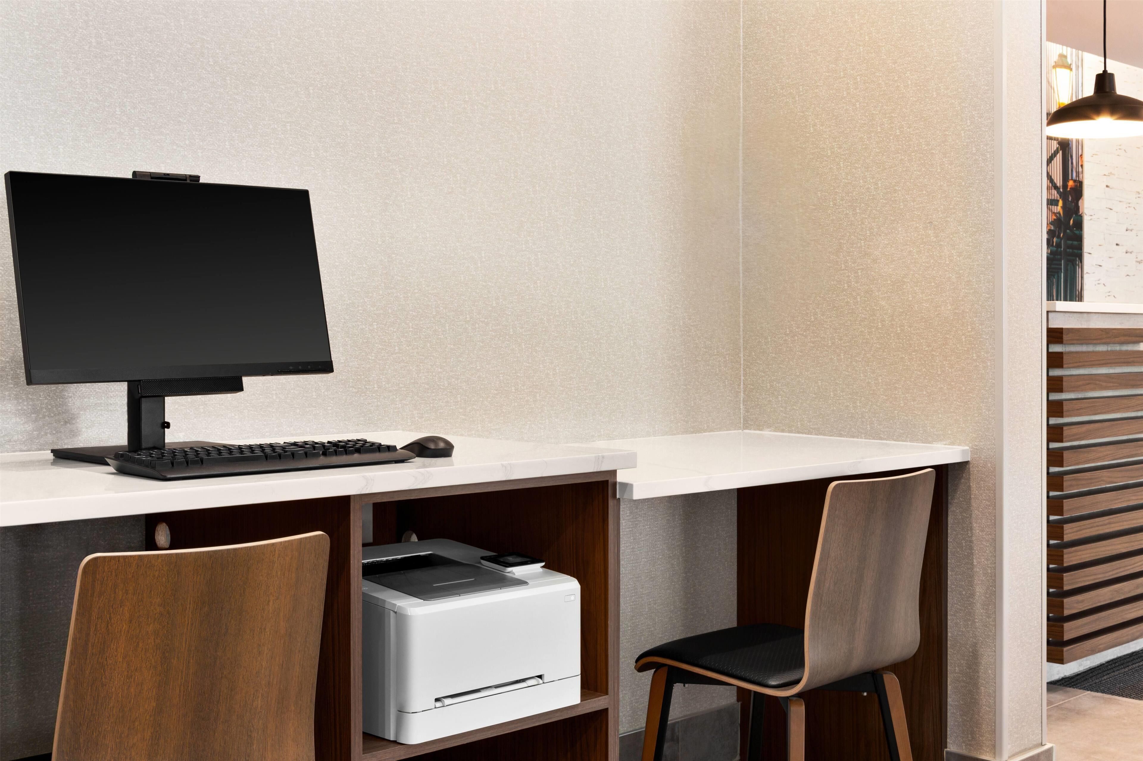Een moderne werkplek met een flatscreen monitor, toetsenbord en printer op een wit bureau. Er staan twee stoelen met houten frames en zwarte kussens.