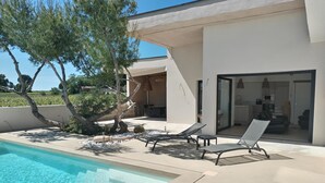 A modern outdoor pool area with lounge chairs, a tree, and a view of the kitchen through glass doors. - Air-conditioned villa, heated pool, vineyard view, calm setting (Pont-Saint-Esprit)