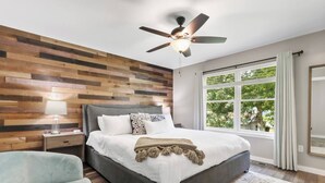 Quarto com cabeceira de madeira, cama com roupa de cama branca, ventoinha no teto, mesa de cabeceira com abajur, janela com cortinas e uma cadeira.