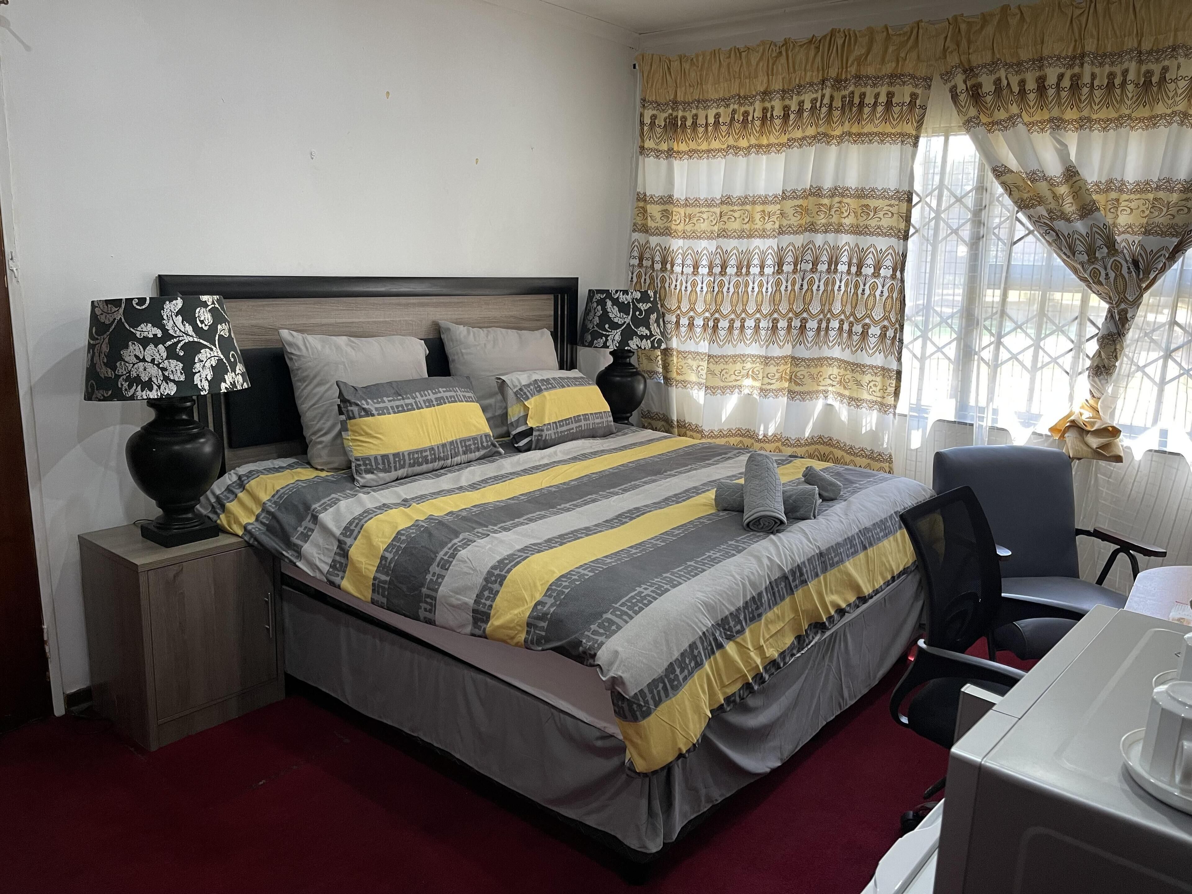 Cama bem arrumada com roupa de cama listrada, mesa de cabeceira com abajur e janela com cortinas estampadas.