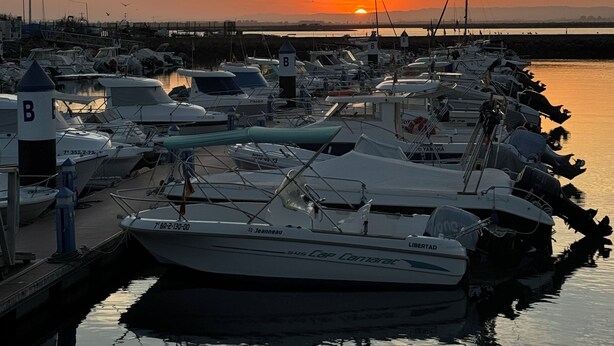 A marina with boats docked at the pier, a sunset in the background, and calm water reflecting the boats.