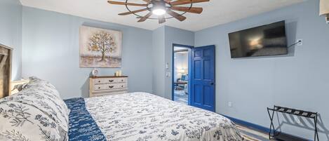 A bedroom with a bed, a ceiling fan, a painting, a TV, and a door.