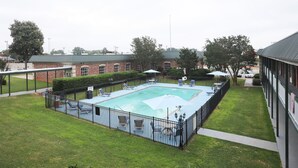 Una piscina rectangular con valla metálica negra, rodeada por un césped y un edificio de ladrillo al fondo.