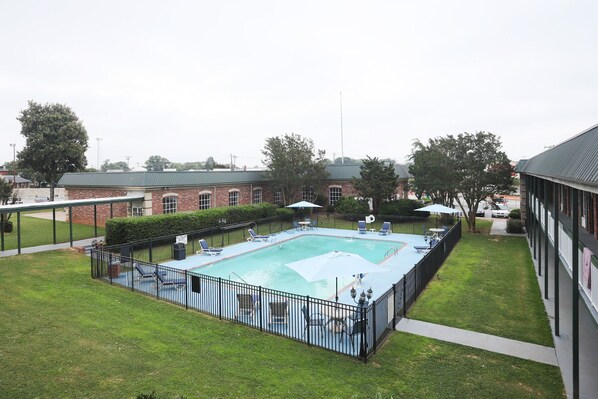 A rectangular swimming pool with a black metal fence, surrounded by a grassy lawn and a brick building in the background. - Greenville Inn and Suites (Greenville)