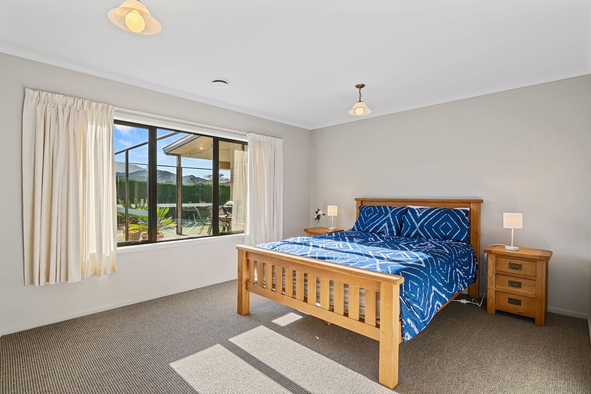 A bedroom with a wooden bed, two bedside tables, a window with curtains, and a view of greenery outside.