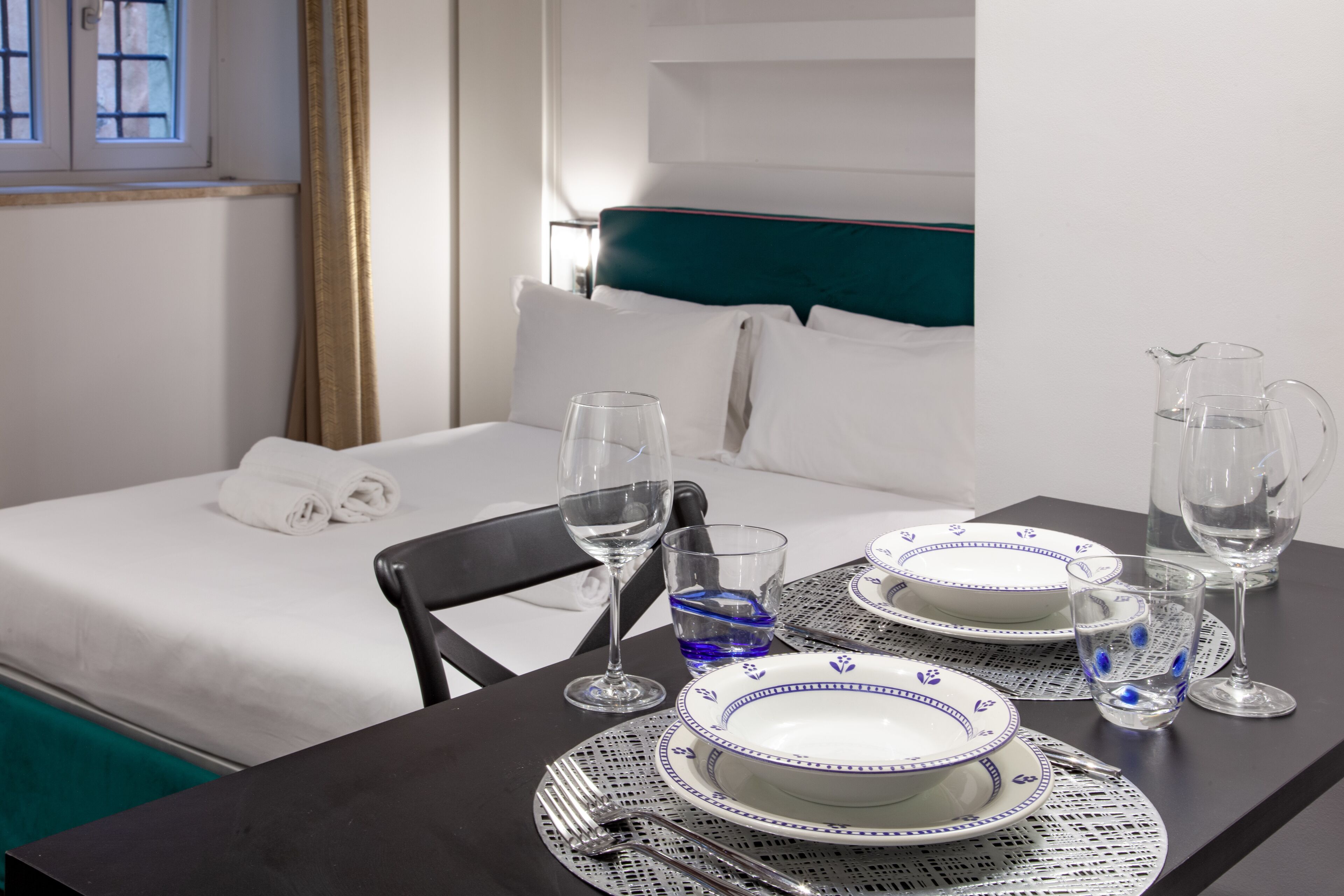 A neatly made bed with white linens and a teal headboard, a dining table set with plates, glasses, and cutlery, and a window with curtains.