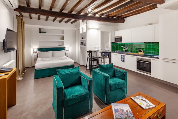 Ein modernes Hotelzimmer mit einem Bett, zwei grünen Sessel, einem Holztisch, einem Schreibtisch und einer Küchenzeile mit grüner Spritzschutzwand.