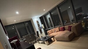 A modern living room with a sofa, a dining table, and chairs. Large windows offer a view of the city at night. - Stunning View 2 Bed Apartment - Canary Wharf City (London)