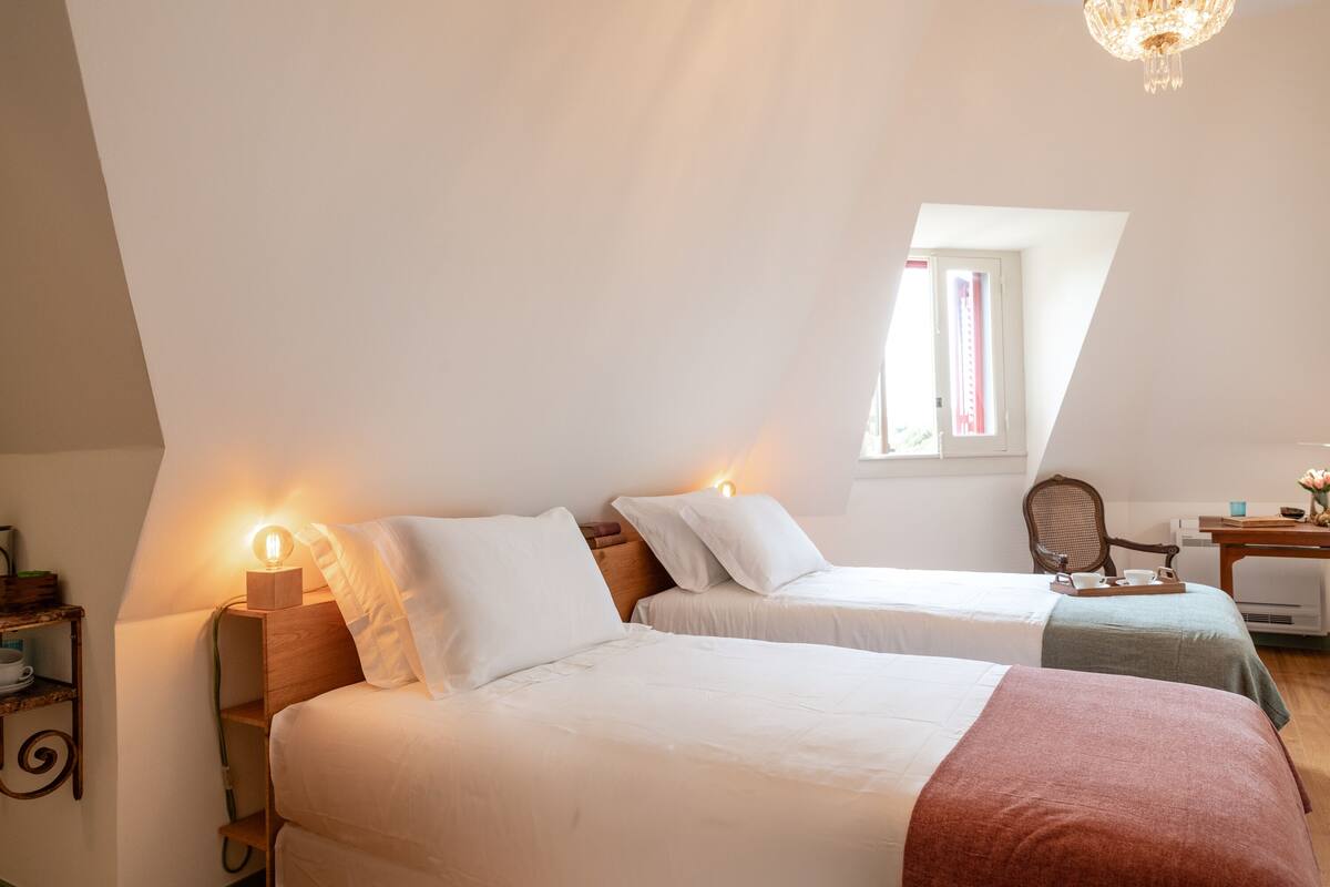 A bedroom with two beds, a wooden nightstand, a chair, a table with a tray, and a window with curtains.