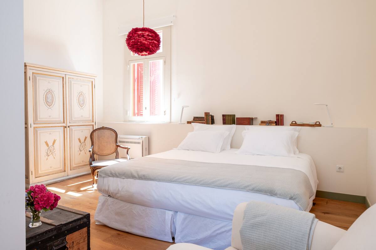A bedroom with a large bed, a dresser, a chair, a window with curtains, and a hanging red spherical light fixture.
