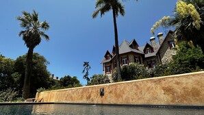 A swimming pool with clear water, surrounded by a stone wall and palm trees. In the background, there is a large stone building with red window shutters. - Chalet Ficalho (Cascais)