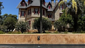 A large stone house with multiple chimneys, red shutters, and a fountain in the foreground. - Chalet Ficalho (Cascais)
