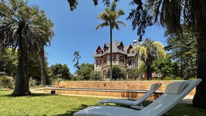 A large stone house with a stone facade and red window shutters, surrounded by lush greenery and palm trees. - Chalet Ficalho (Cascais)