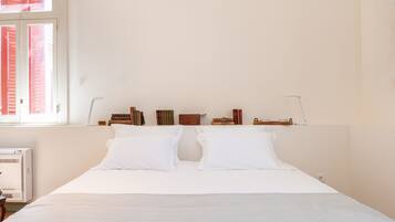 A neatly made bed with white linens, a bedside table with a lamp, and a window with closed shutters.