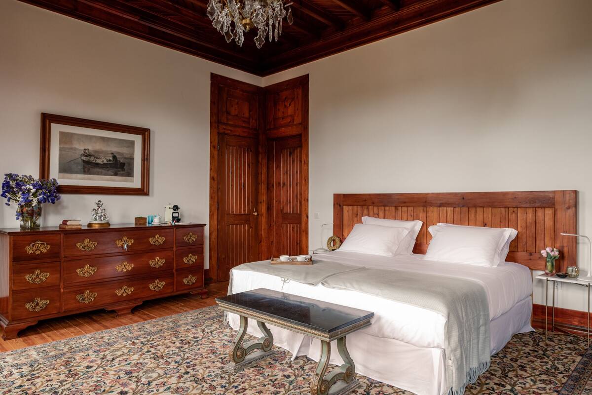 A bedroom with a wooden headboard, a large bed, a nightstand, a dresser with drawers, a framed picture on the wall, and a chandelier.