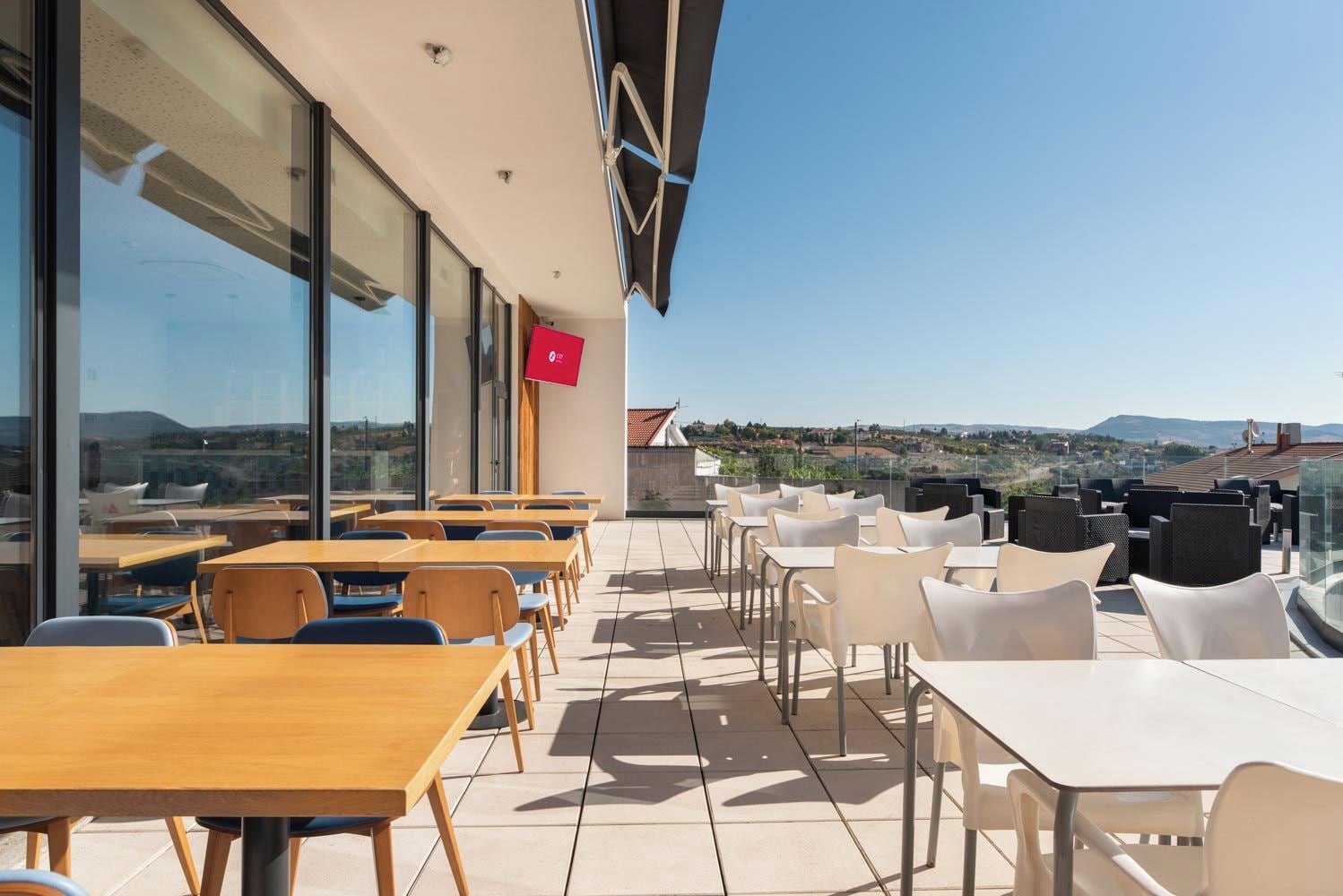 Una terraza exterior con mesas y sillas, amplias ventanas y vistas del paisaje.