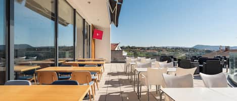 En udendørs terrasse med borde og stole, store vinduer og udsigt over landskabet.
