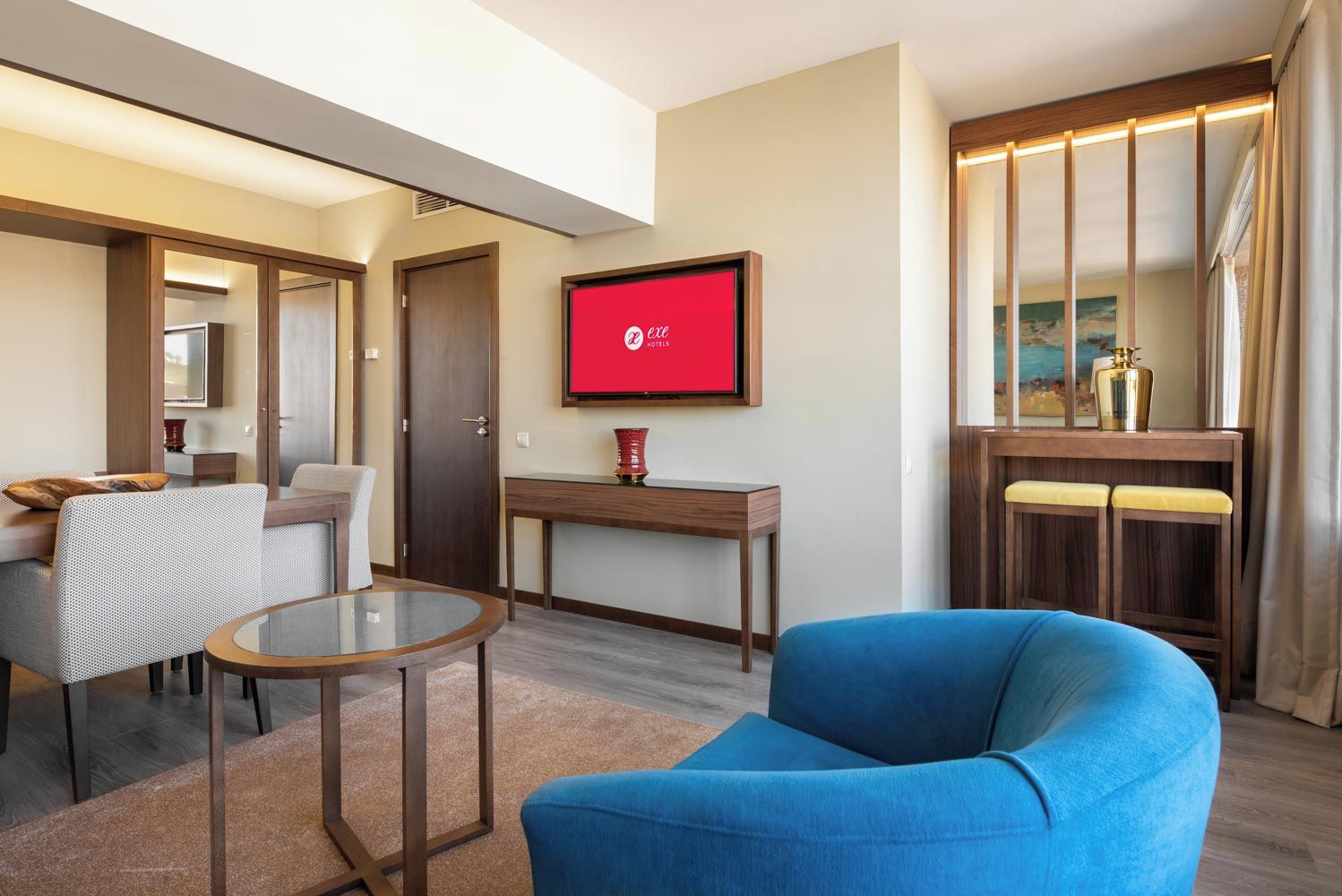 Een moderne hotelkamer met een blauwe fauteuil, een glazen salontafel, een houten bureau en een televisie waarop het logo van het hotel staat.