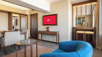 Een moderne hotelkamer met een blauwe fauteuil, een glazen salontafel, een houten bureau en een televisie waarop het logo van het hotel staat.