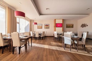 A spacious dining area with multiple tables and chairs, large windows with curtains, and artwork on the walls.