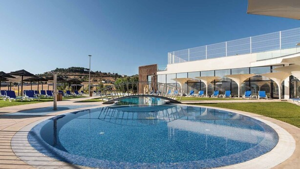 En stor udendørs swimmingpool med cirkelformet design, omgivet af liggestole og parasoller, med en moderne bygning i baggrunden.